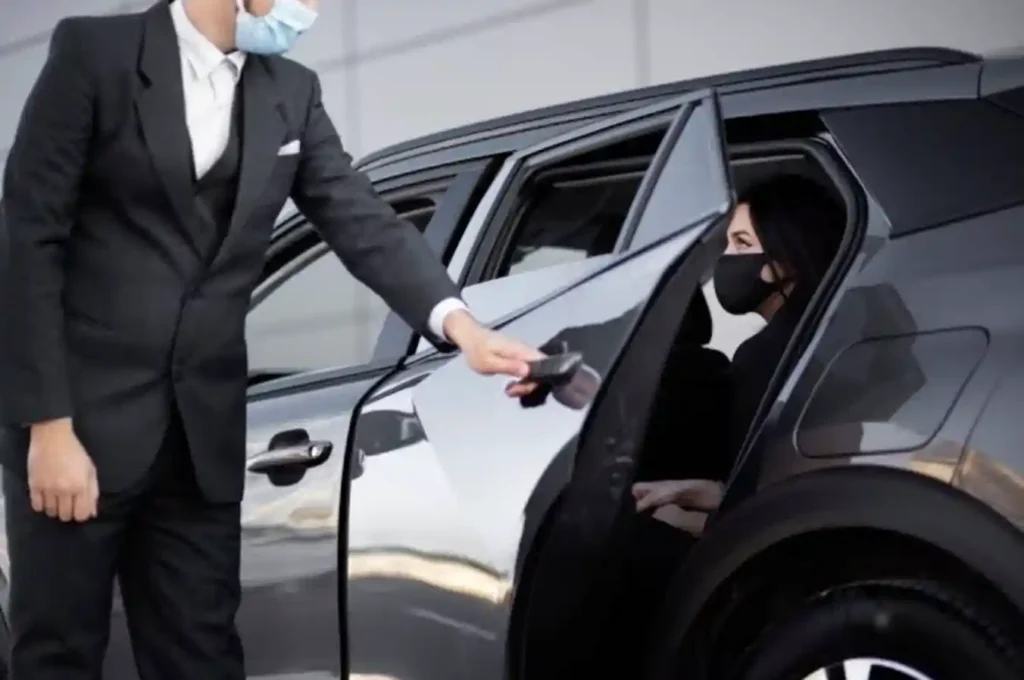 A professional chauffeur in a suit opens the car door for a female client wearing a face mask, representing a VIP or private transfer service with safety precautions.