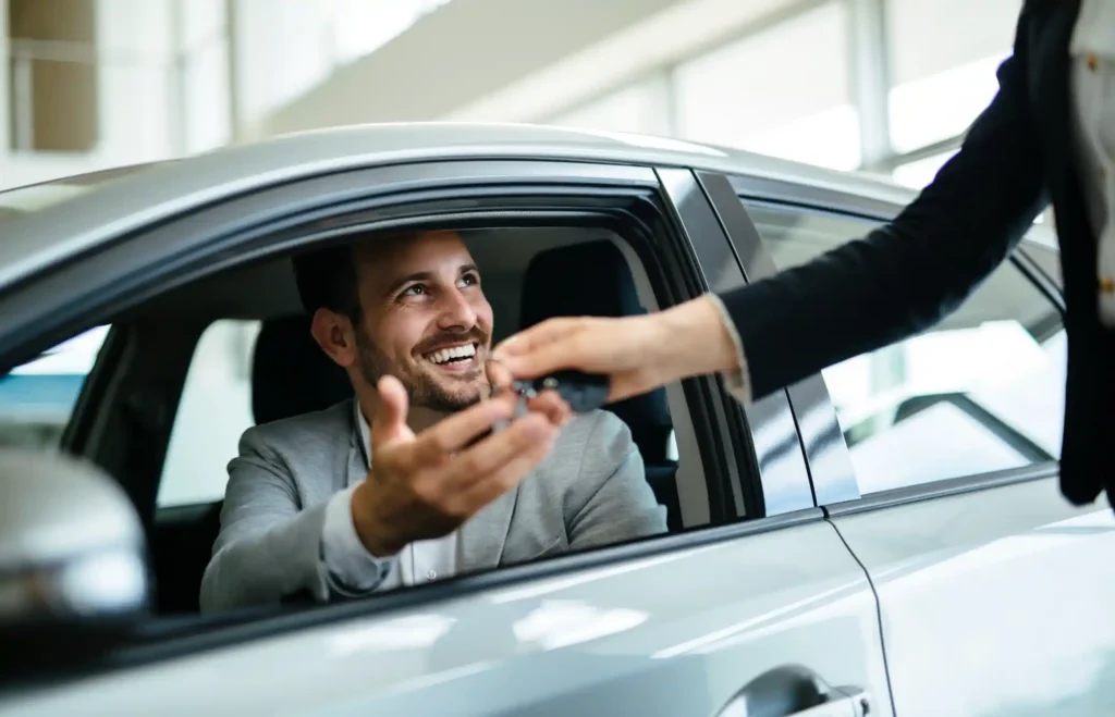 A smiling man sitting in a car receives the car keys from a hand extended through the window, representing a car rental handover or vehicle delivery moment.